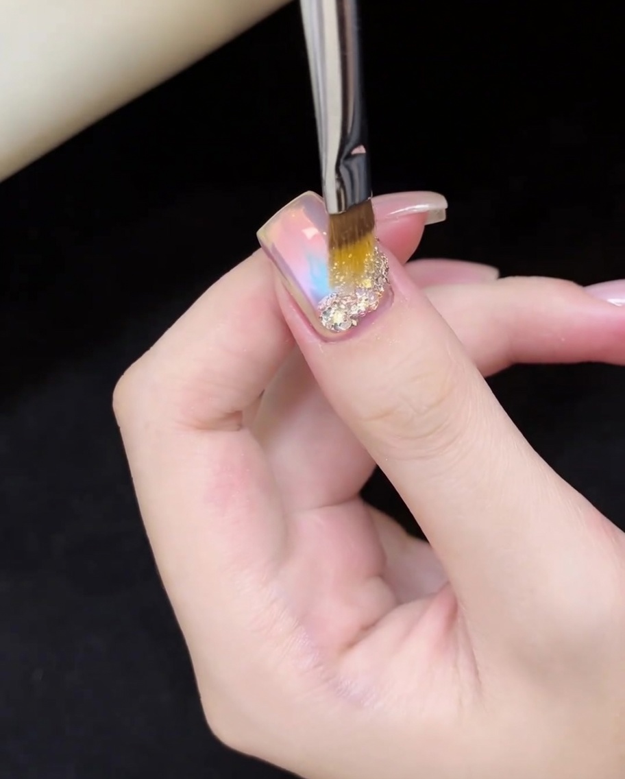A nail brush precisely placing shimmering, iridescent gold and silver glitter flakes onto the base of the nail.