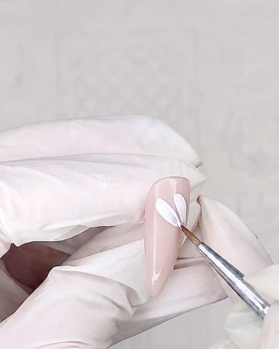 A detail brush painting white petals onto a pink-coated nail.