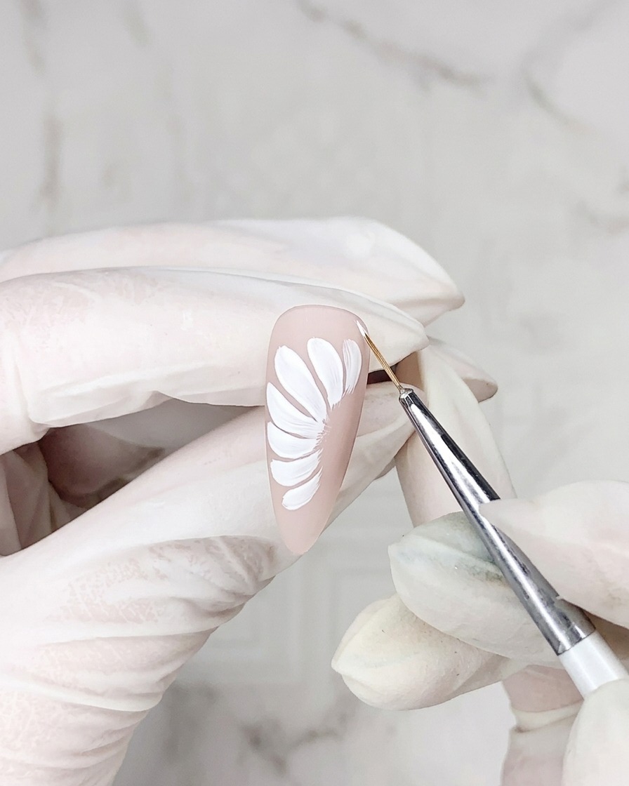 A close-up view of a detail brush applying a thin line of adhesive gel around the edge of a white daisy petal painted on a nude nail base.