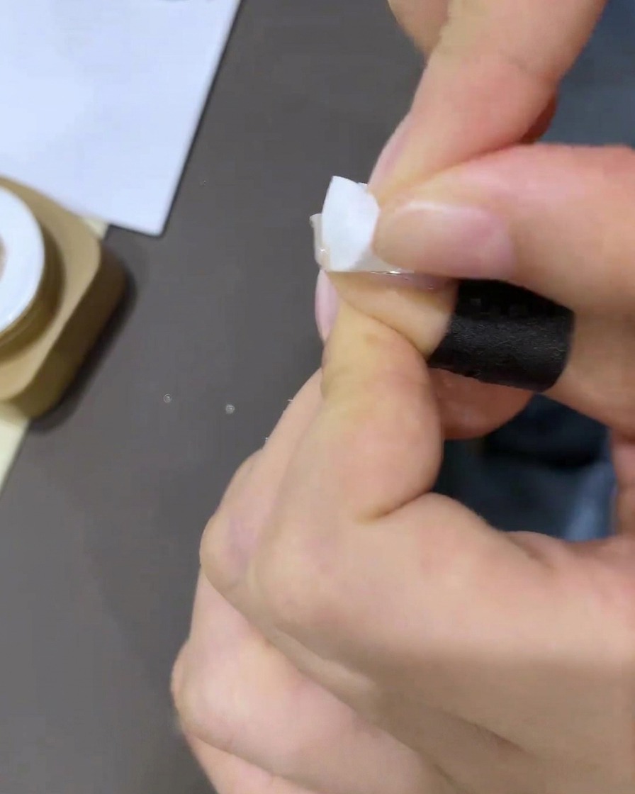 Close-up of a hand applying a small piece of white concealer tape to the base of the nail cuticle.
