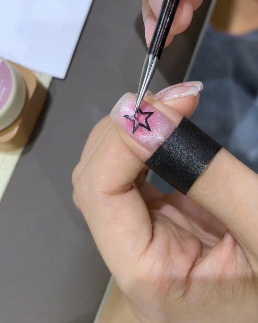 A close-up showing a pair of tweezers placing a small four-pointed star sticker onto the nail surface.