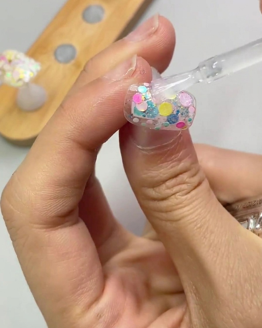 A brush applying a clear layer of reinforcement gel over a glitter-decorated nail.