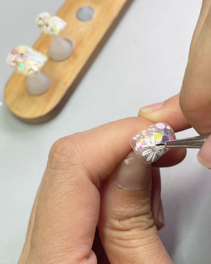 A close-up view of a brush using white sculpting gel to form the petals of a second small daisy on the nail surface.