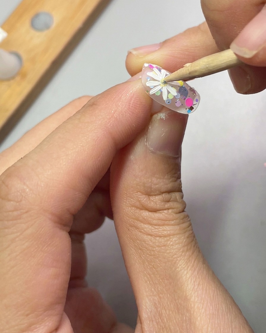 A dotting tool placing a small drop of yellow gel into the center of a 3D white daisy on a decorated nail.