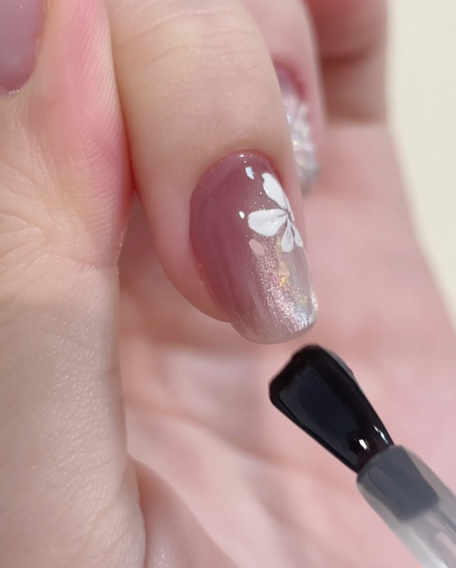 A gel brush applying a clear, glossy top coat over a finished nail design featuring cat-eye gel, glitter, and white floral art.