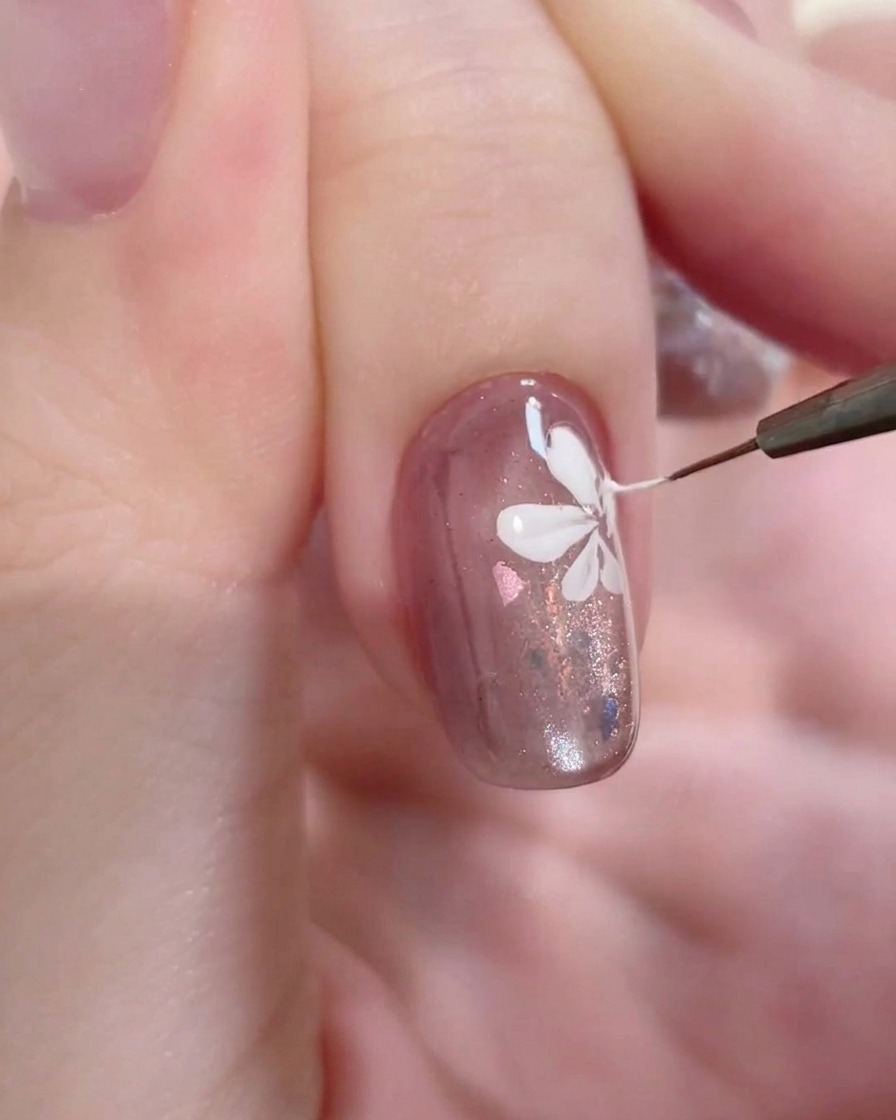 A macro shot of a fine liner brush applying a precise white stroke to the center of a painted flower on a gel-coated nail.