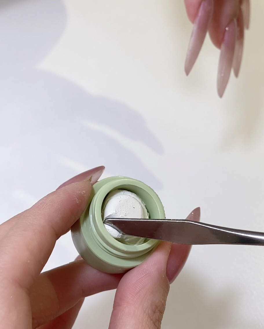 A close-up of a metal spatula picking up a small amount of ivory white gel from its container.