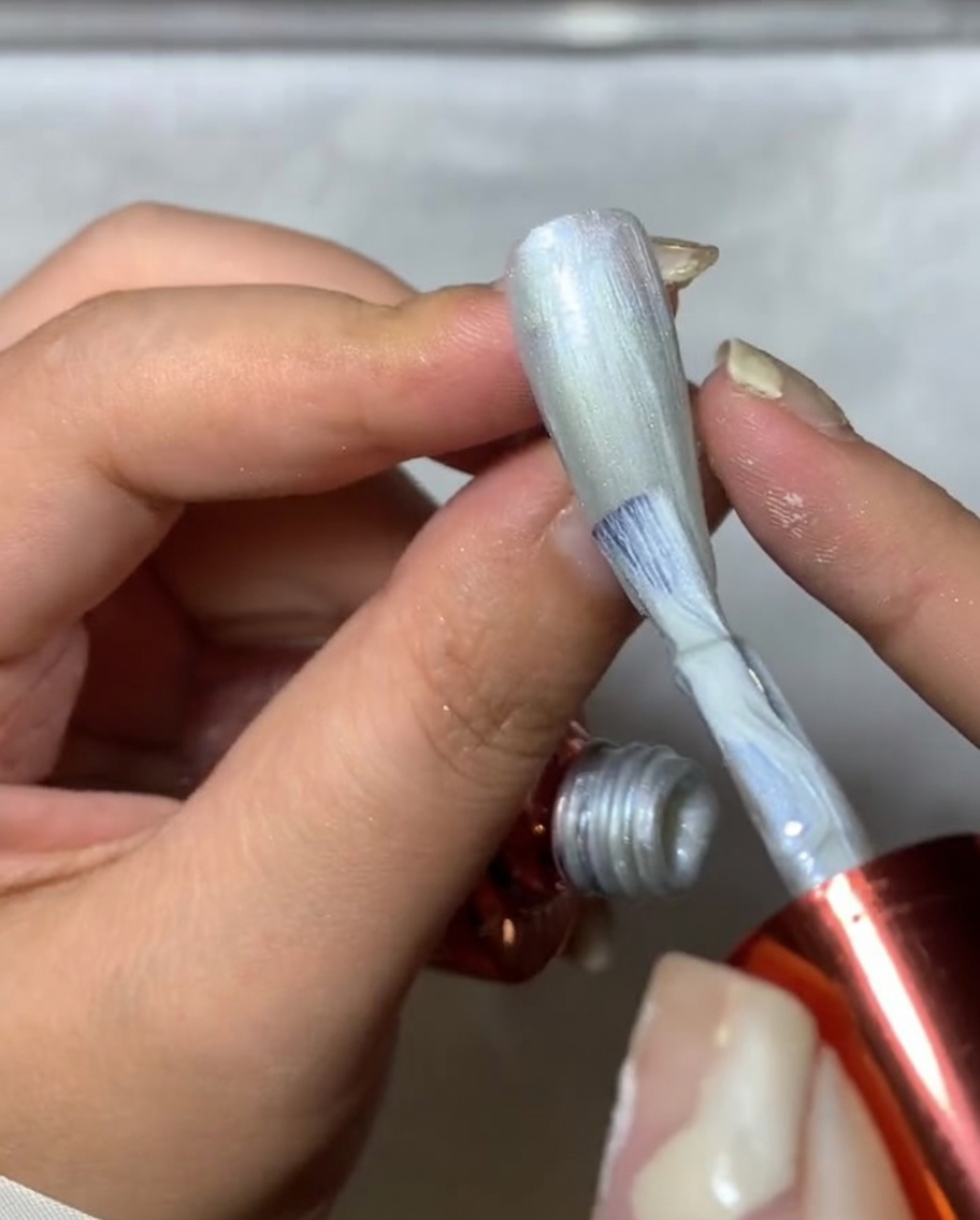A close-up of a nail brush applying silver spider gel onto a nail, creating fine, textured, web-like lines.