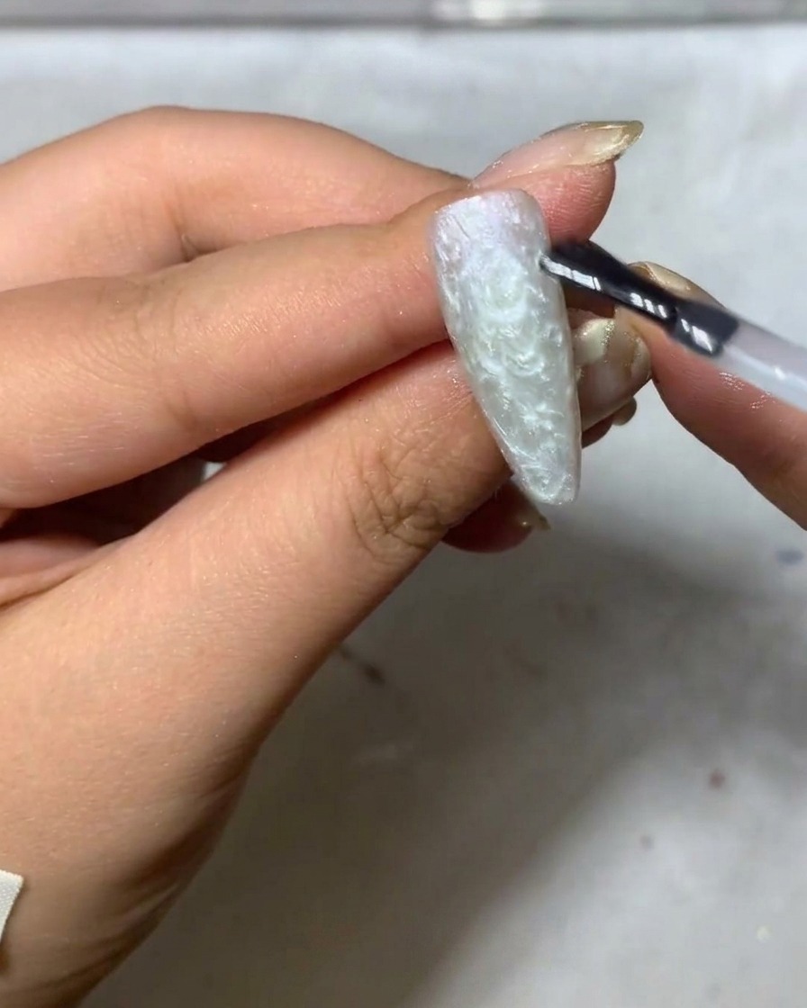 A fine liner brush beginning to paint delicate white daisy petals onto a matte grey-toned nail surface.
