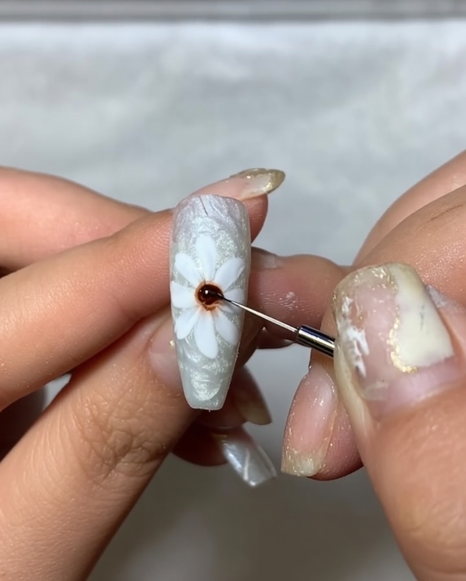 A close-up of a nail with painted white petals, using a fine brush to apply a dot of dark brown gel to the center.