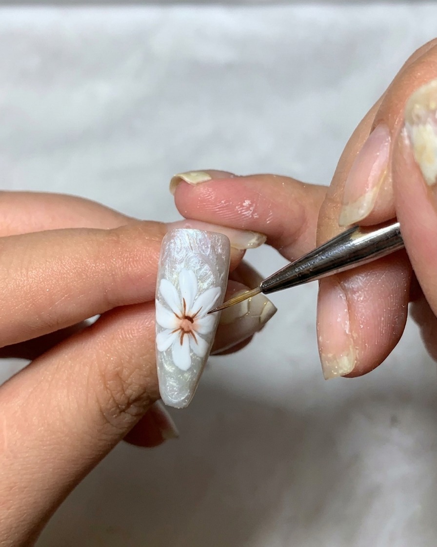 A fine brush applying a thick, clear gel over the white petal design on the nail to build dimension.