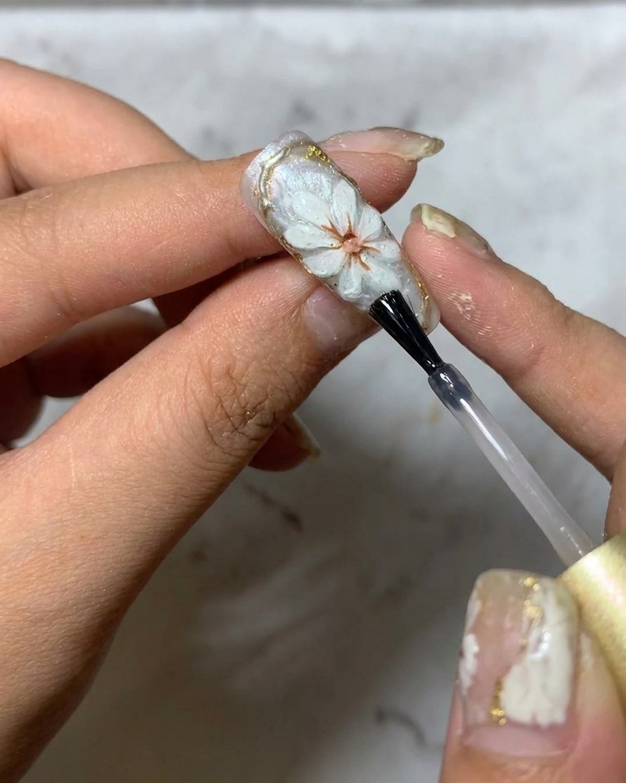 A brush applying a final glossy top coat over a 3D daisy nail art design.