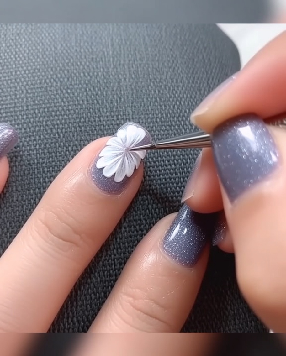A close-up shot of a fine liner brush applying a small dot of black gel paint to the center of a painted white daisy on a light gray nail base.