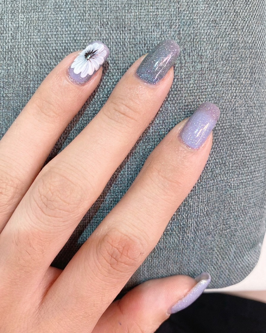 A hand applying a thin layer of top coat over a finished nail design featuring a white daisy and a small text sticker.