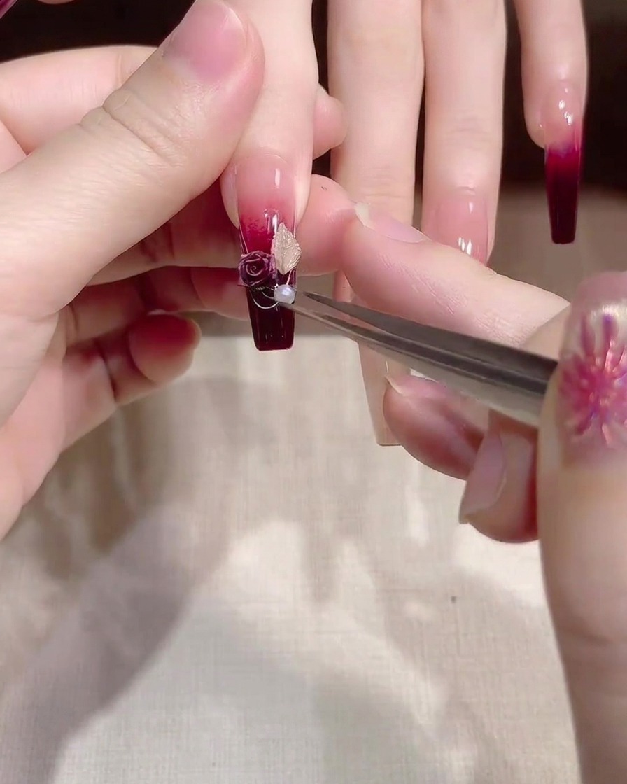 Tweezers placing a small, shiny white pearl onto the center of a floral charm on a nail.