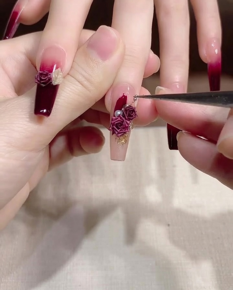 A thin detail brush being used to nudge and adjust small gold and pearl decorations on a fingernail.
