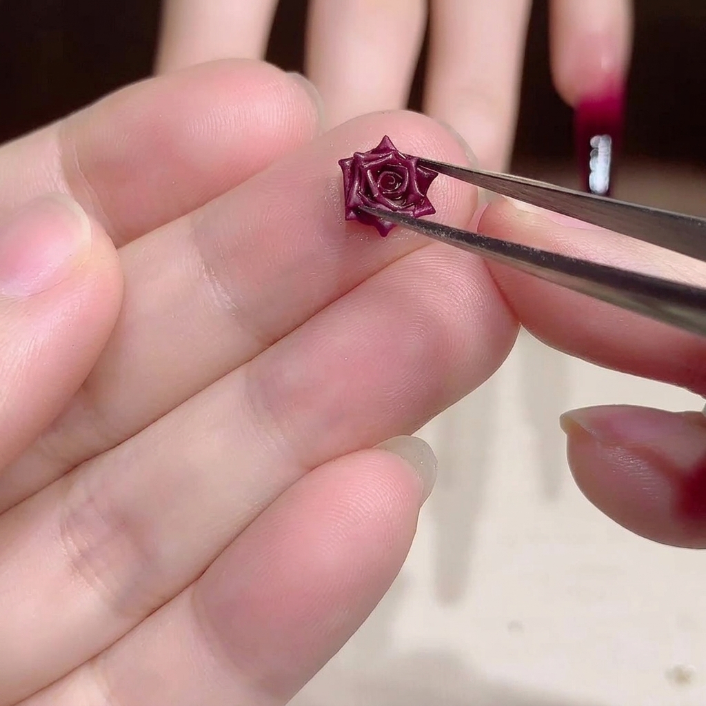 Close-up of a pair of metal tweezers holding a small, intricate, dark red 3D dried rose nail art charm.