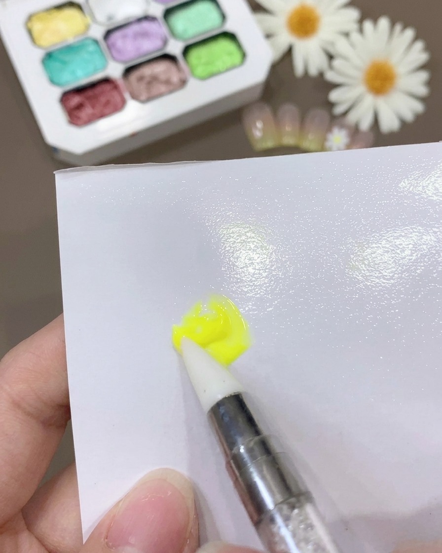 A brush blending yellow gel and base coat on a white surface to create a thin, consistent mixture.