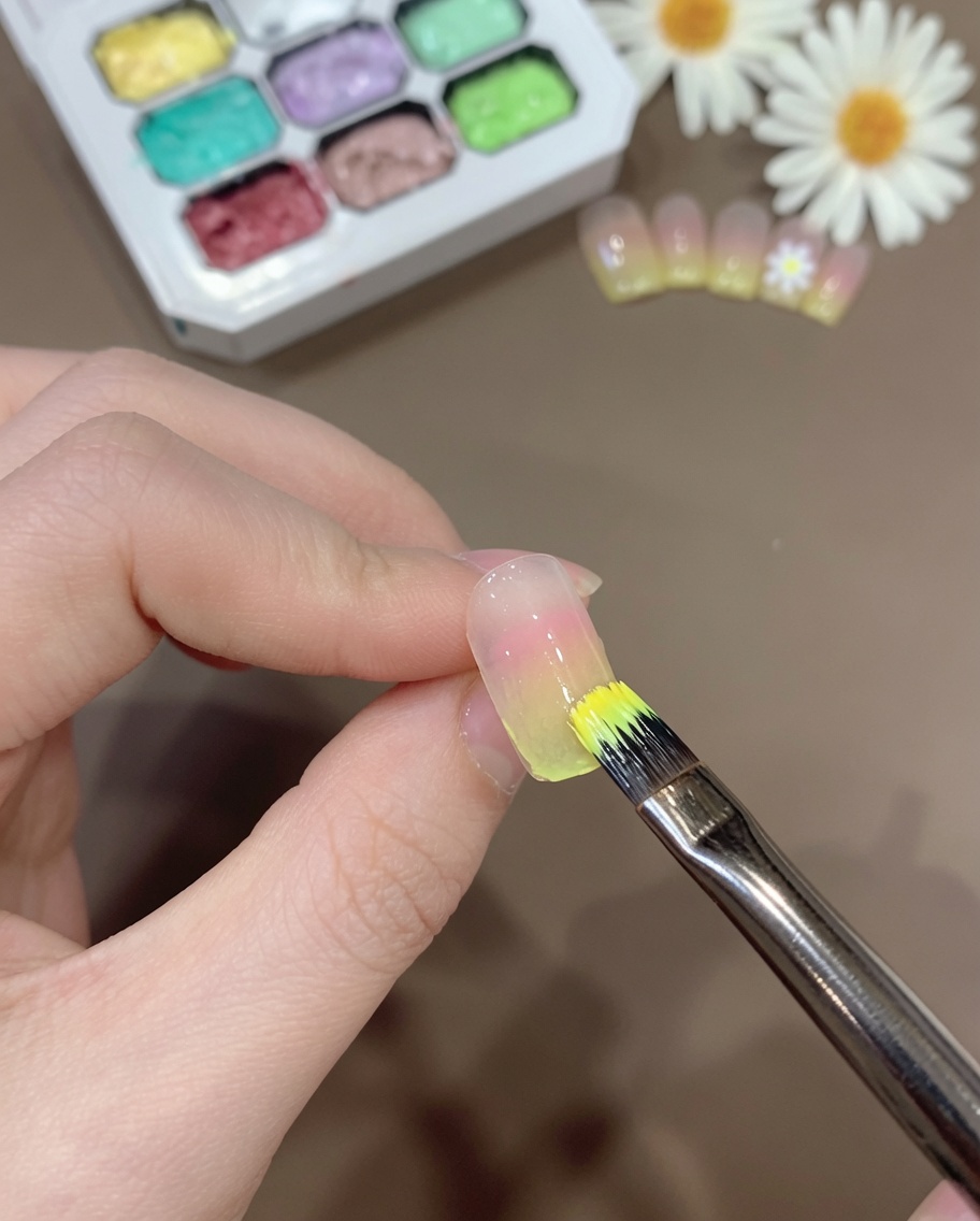 A brush applying a thin layer of sheer yellow gel mixture onto the tip of a gradient-colored nail.