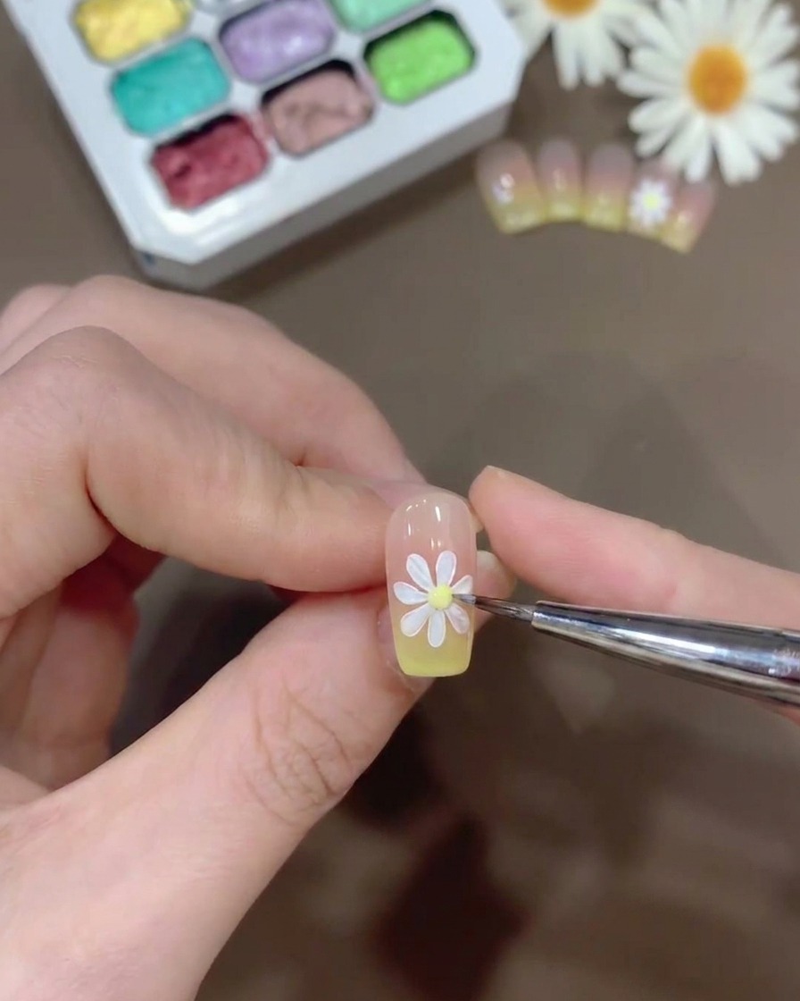 A tiny bead of yellow gel being placed in the center of a painted white daisy on a nail.