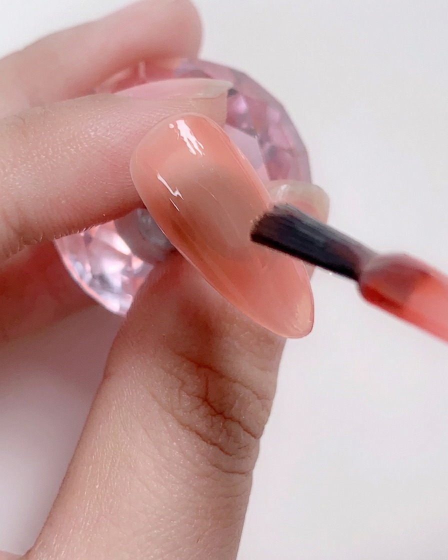A brush applying a coat of soft pink gel polish to the center of the nail.
