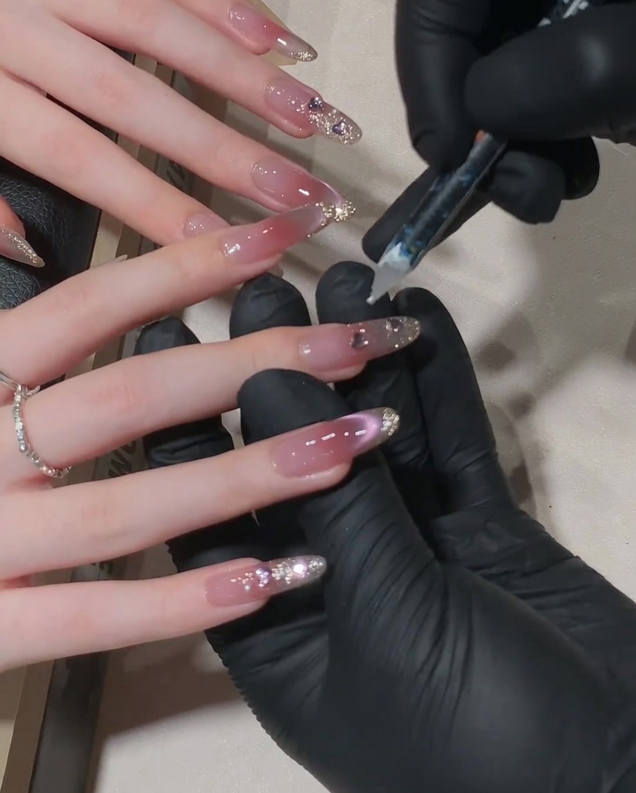 A close-up shot of a nail technician using a wax-tipped tool to place a small crystal at the cuticle line of a pink cat-eye nail.