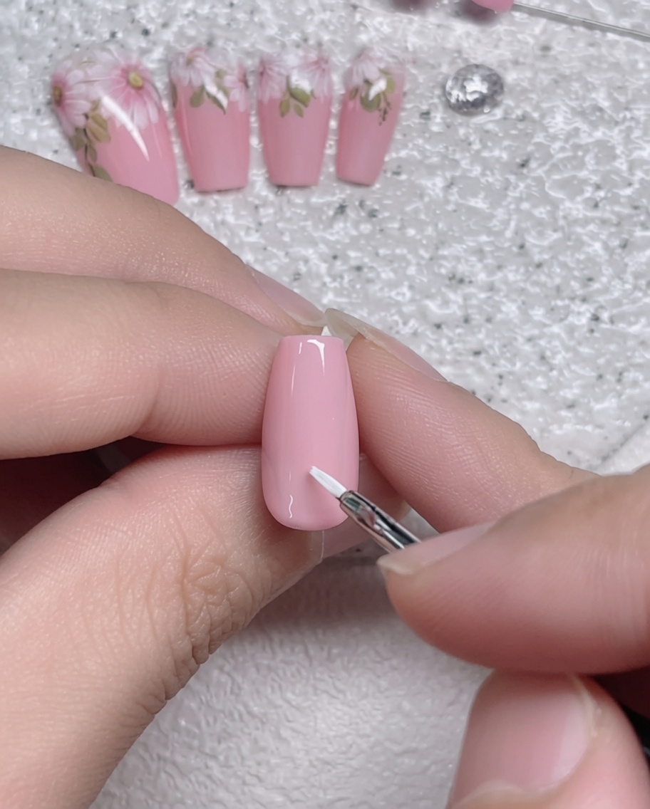 A close-up of a fine liner brush applying a single white petal stroke onto a soft pink gel-coated nail.