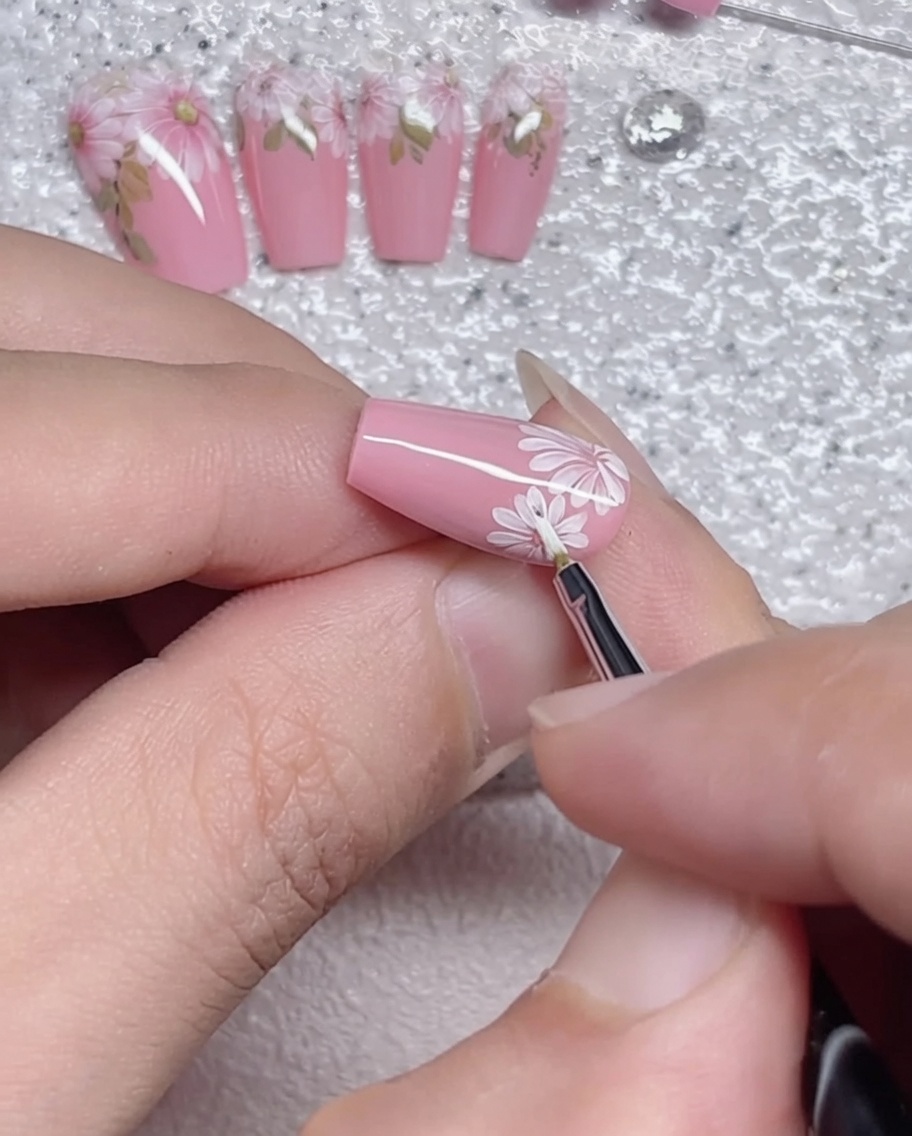 A fine liner brush carefully tracing the perimeter of white daisy petals on a pink nail.