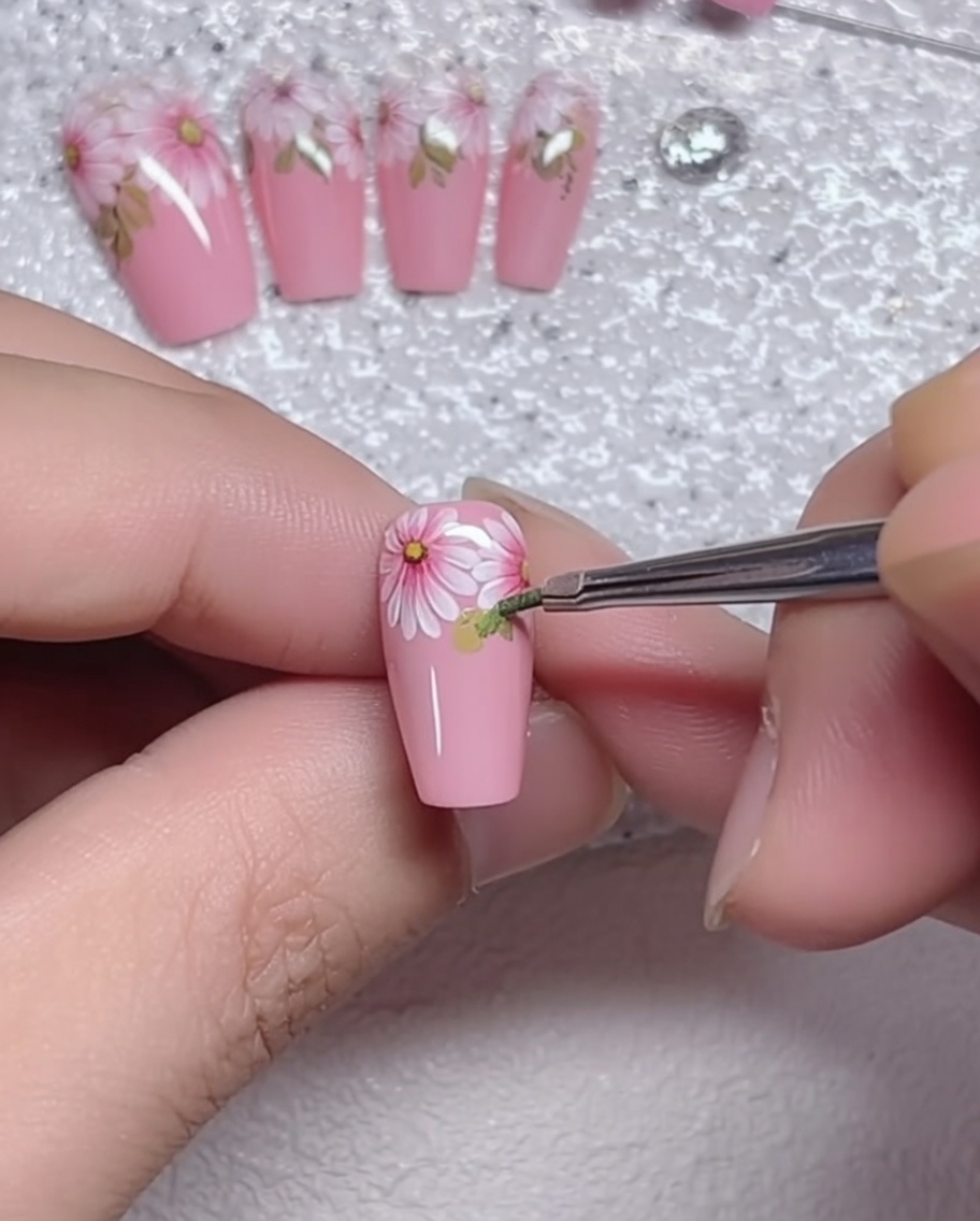 A close-up view of a fine liner brush adding small green leaf details at the base of a hand-painted daisy on a soft pink nail.