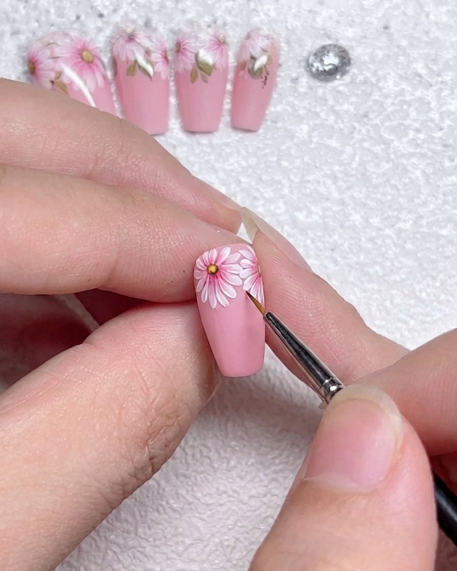 A close-up of a fine liner brush applying precise white highlight lines to the edges of daisy petals on a pink gel-painted nail.