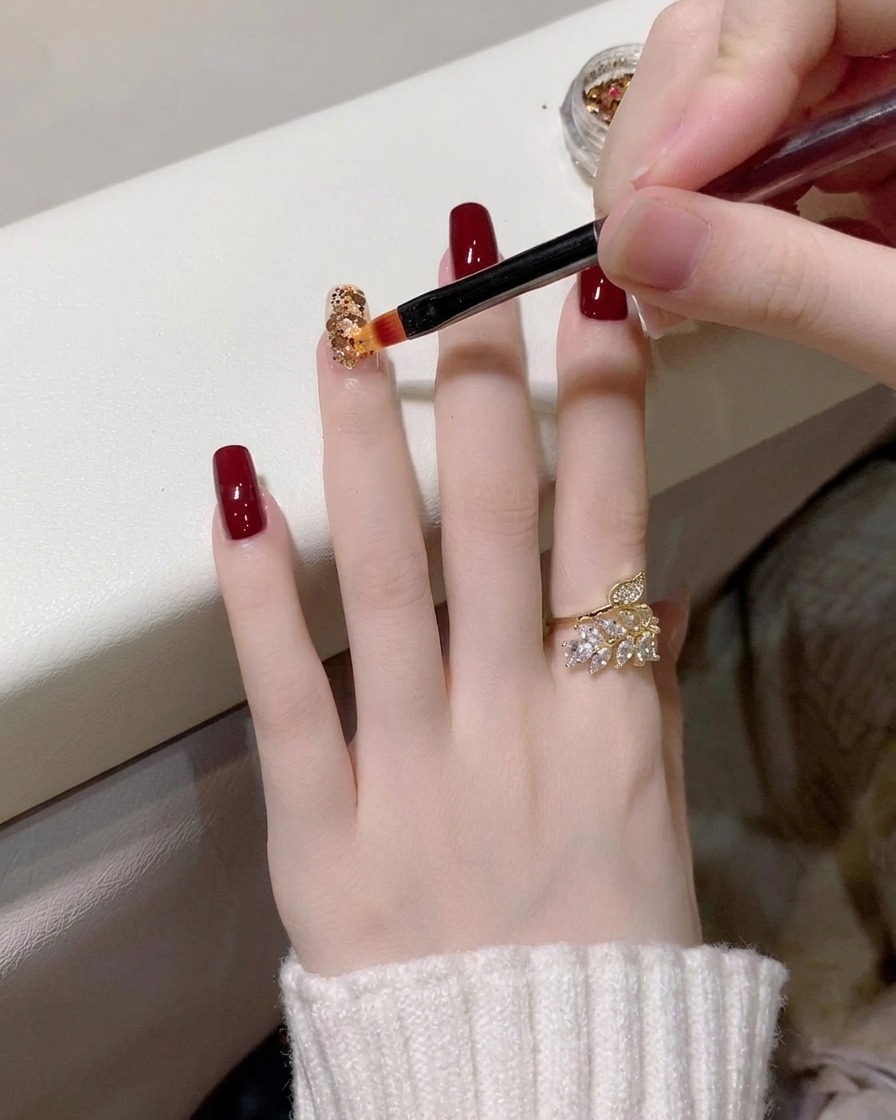 A close-up of a detail brush placing small gold and holographic glitter flakes onto a clear-coated nail.