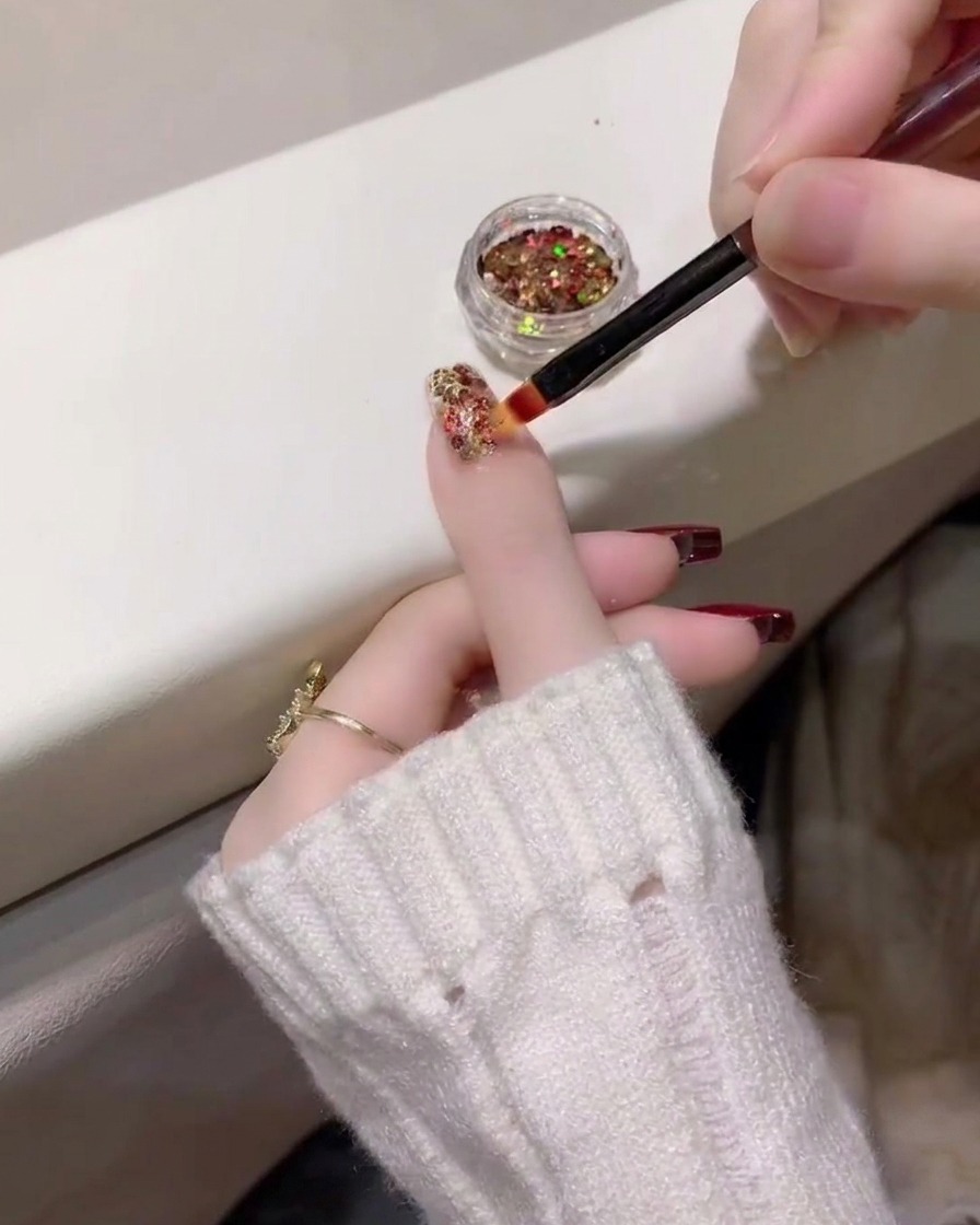 A close-up view showing a dense layer of multi-colored glitter being applied to the ring finger nail.