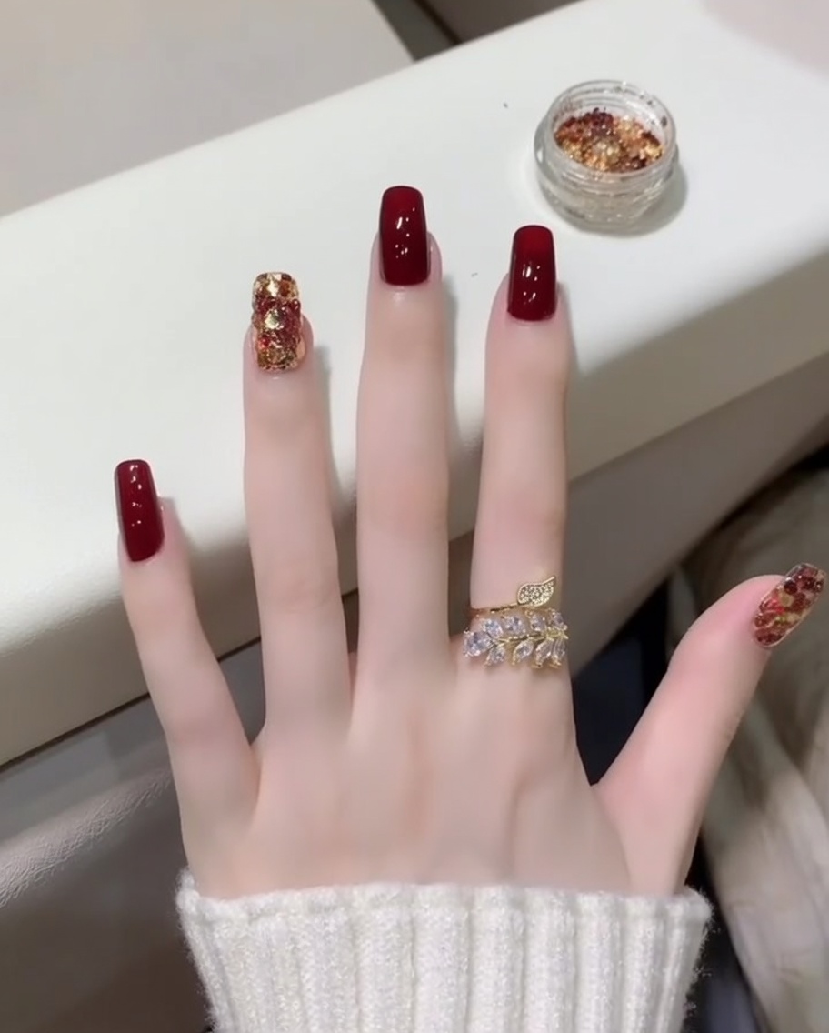 A close-up view of a brush applying a clear, glossy top coat over dark red and glitter-accented nails.