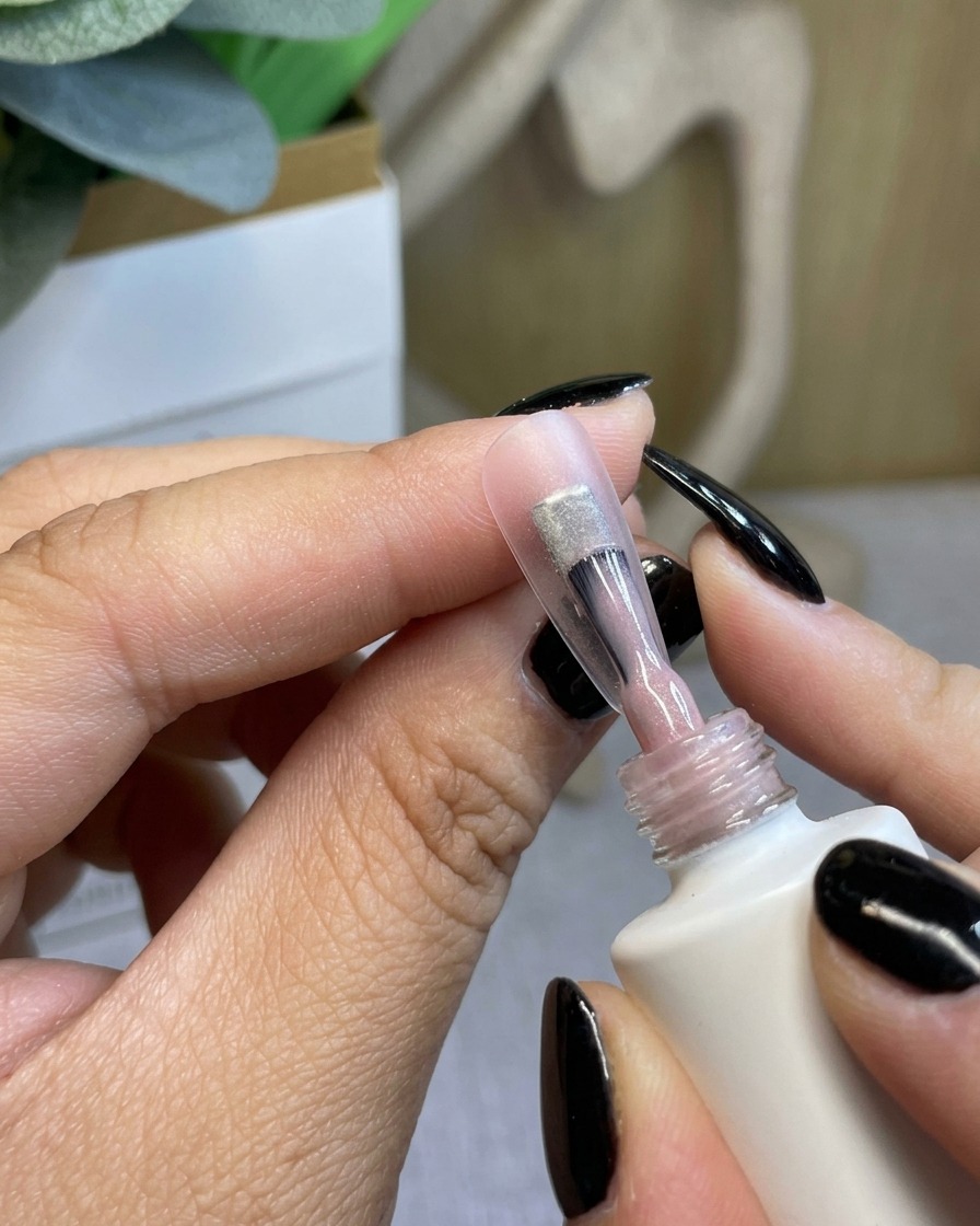 A brush applying a clear, sheer gel base coat to a natural nail.