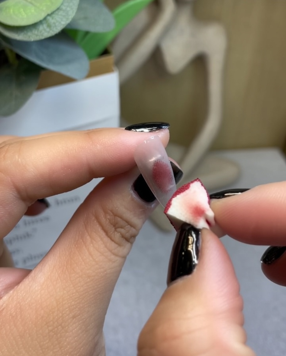 A makeup sponge dabbing deep wine-red gel onto the center of a clear nail tip.