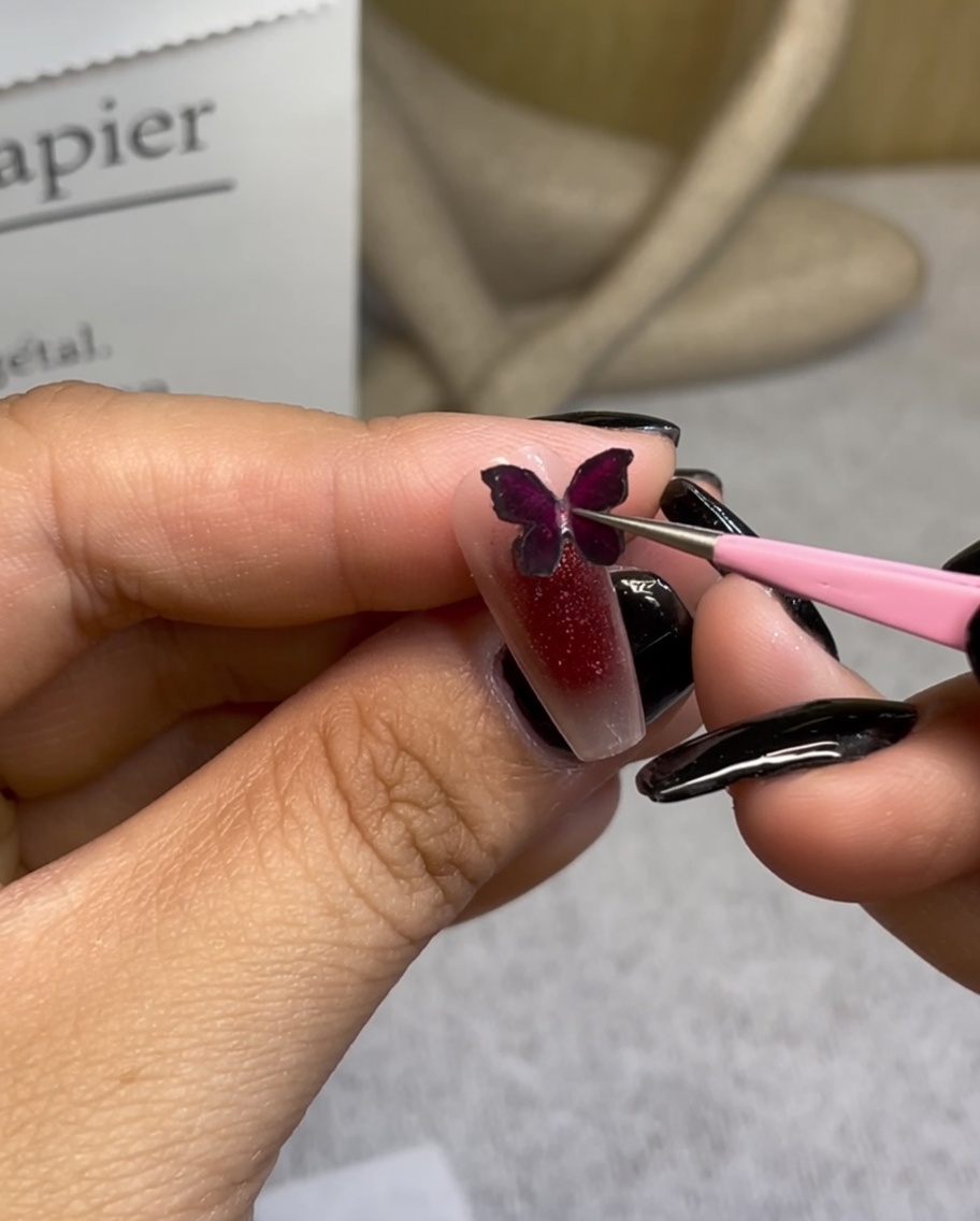 Tweezers placing a decorative burgundy butterfly charm onto a red gradient nail.