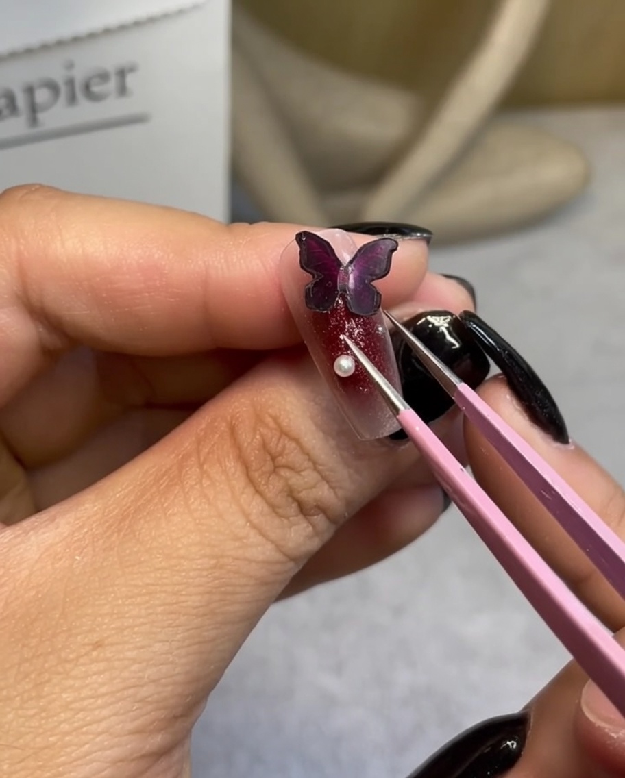 A pair of precision tweezers placing a small white pearl next to a dark butterfly charm on a manicured nail.