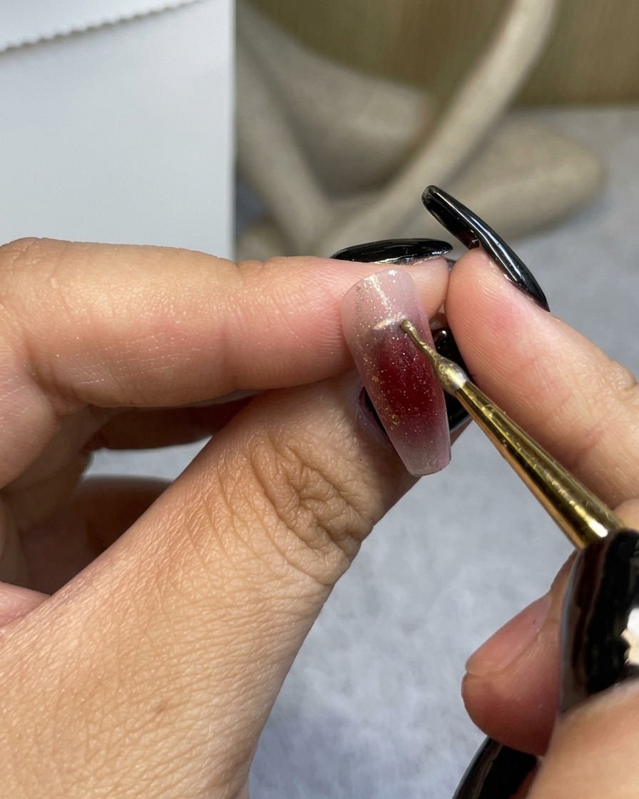 A fine detail brush applying a thin metallic gold line near the cuticle of a wine-red ombre nail.
