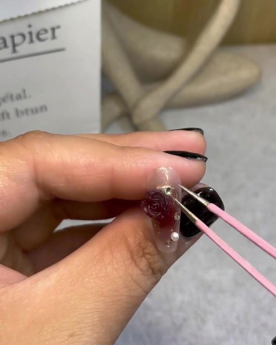 A hand using tweezers to add tiny pearls around a 3D rose on a finished nail design.