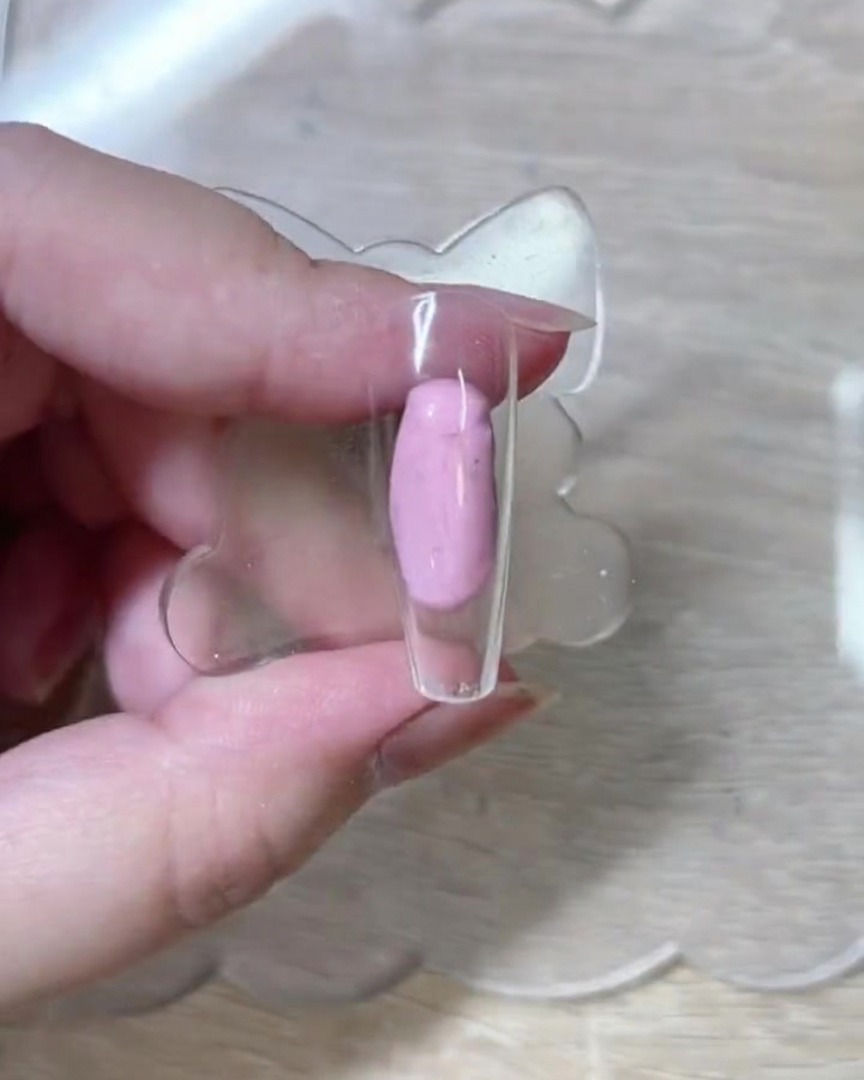 A close-up shot of a fine liner brush drawing delicate white rose outlines on a bare, translucent pink nail tip.