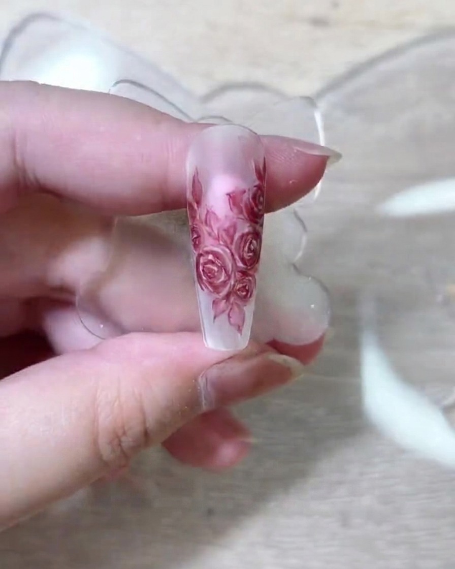 A fine nail art brush depositing deep red pigment onto the layered rose petals on a nude nail extension.