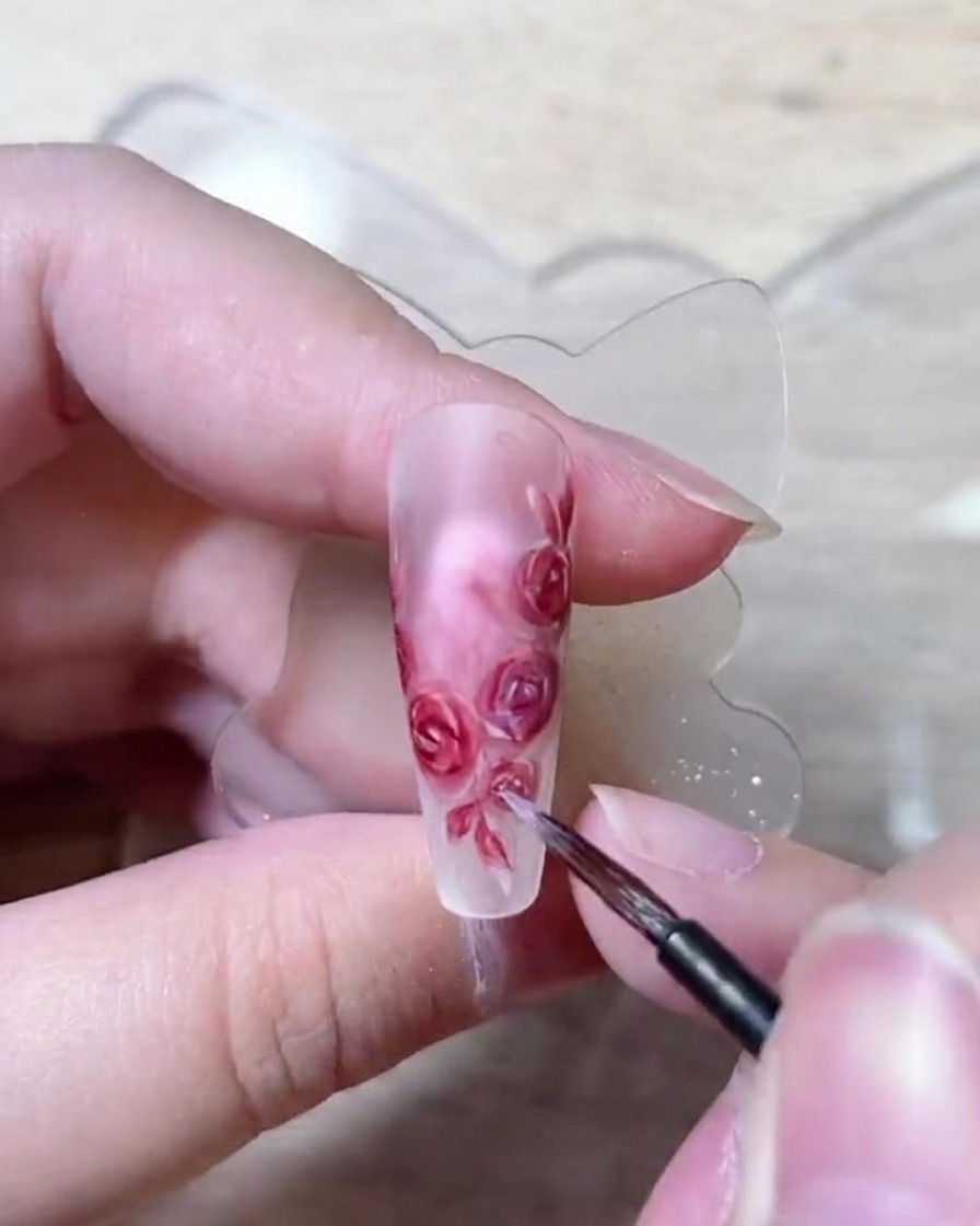 A close-up of a fine nail brush delicately placing white highlight dots onto the edge of painted rose petals.