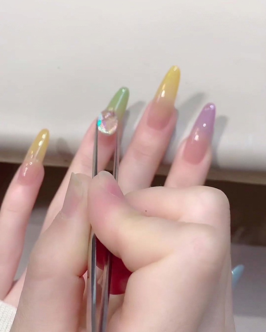A close-up of a pair of tweezers placing a shiny, clear crystal heart-shaped charm onto a jelly-colored nail.