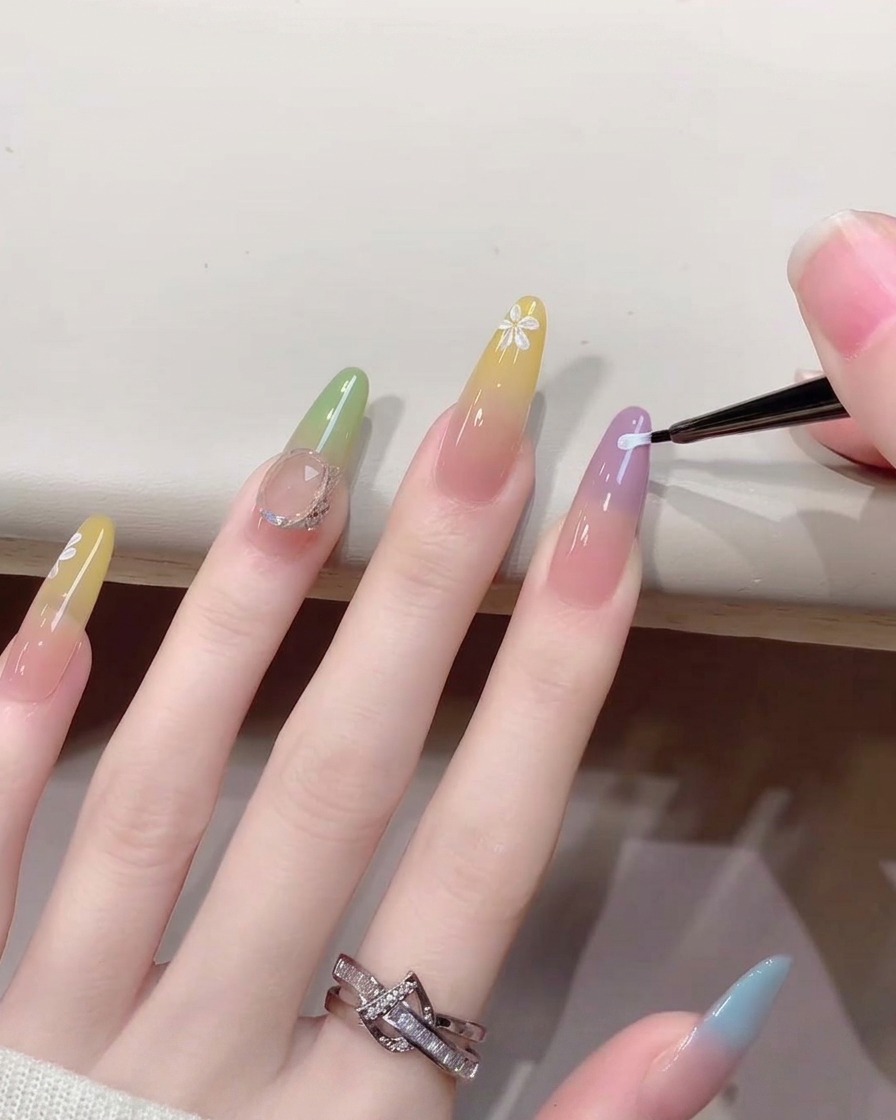A close-up of a fine nail art brush painting tiny white flower petals onto a gradient-colored fingernail.