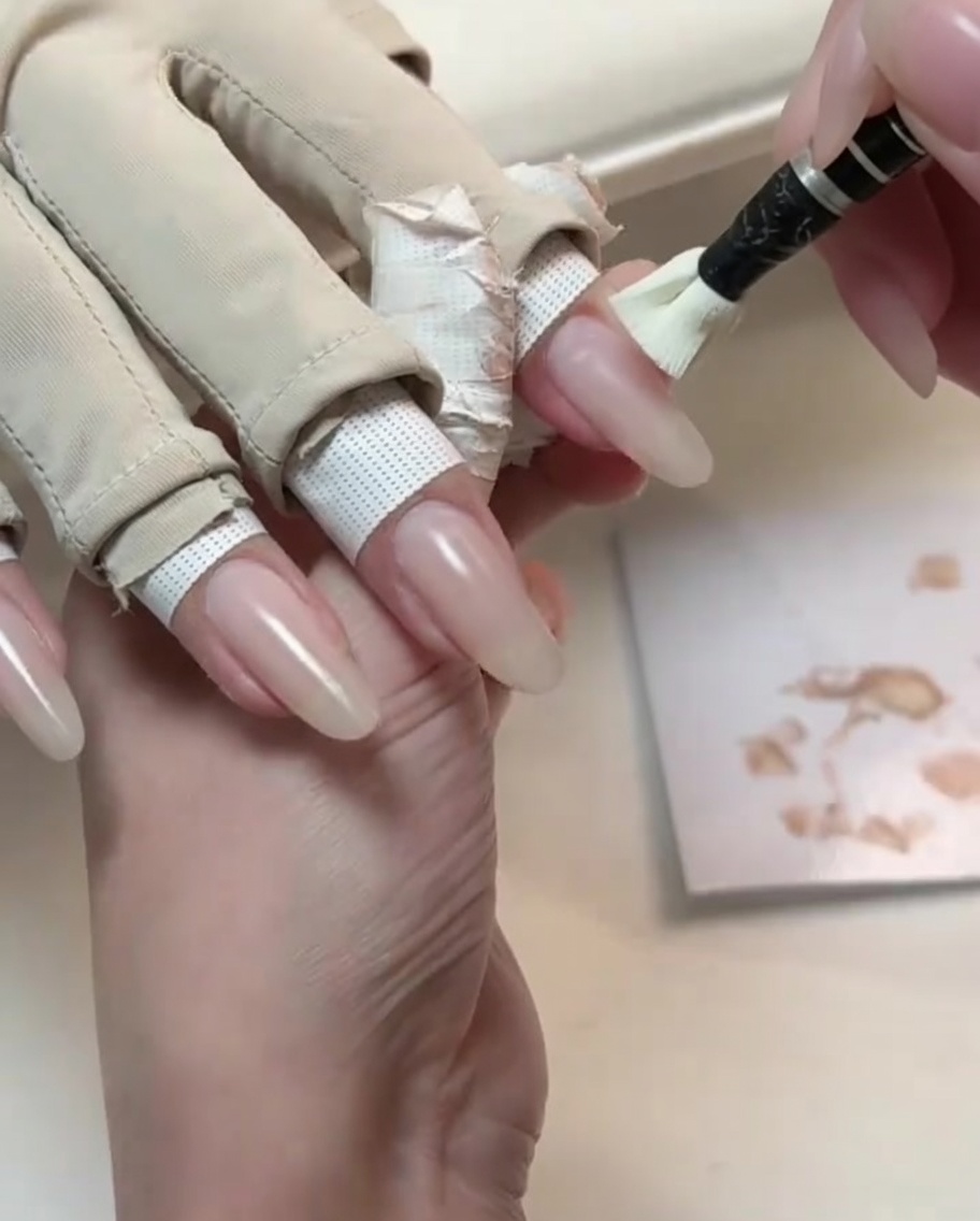 A small application brush coating a long natural-shaped nail with a light nude gel polish.
