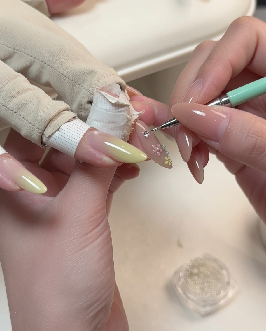 A detailing tool placing a small pearl into the center of a floral nail art design on an almond-shaped nail.