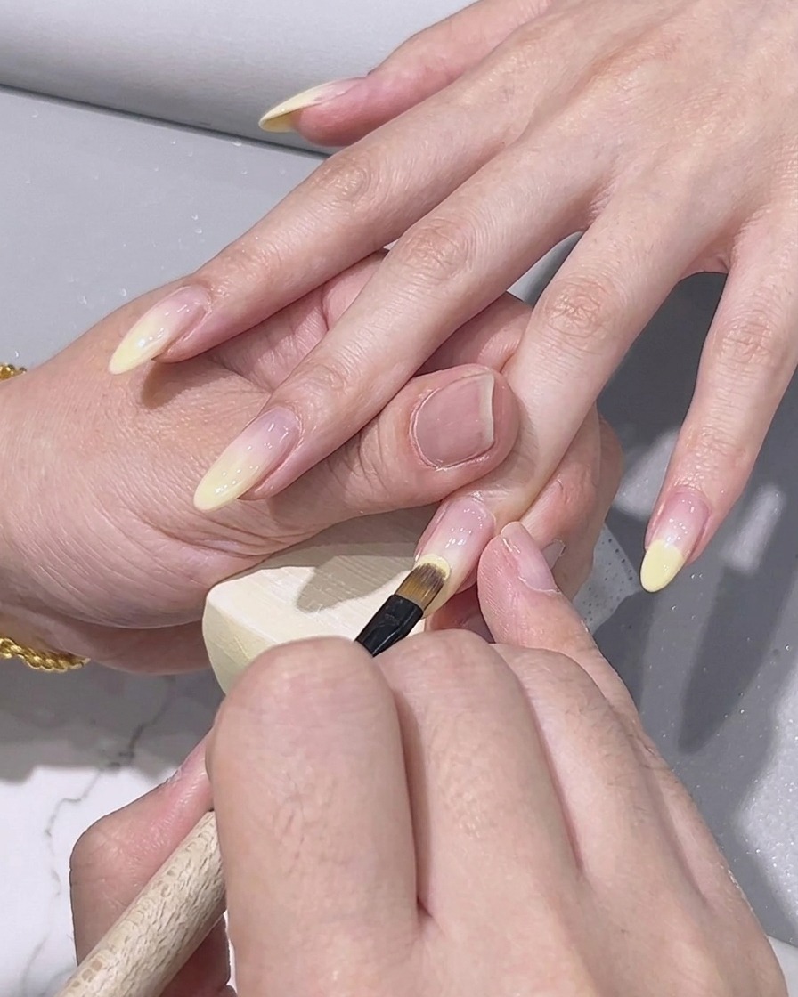 A fine detailing brush applying lemon yellow gel polish onto the tip of a fingernail.