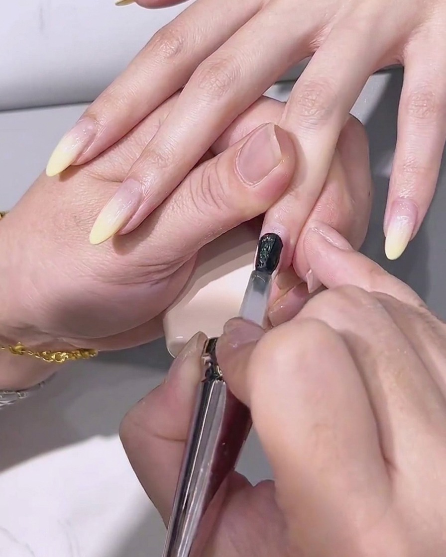 A brush applying a clear, glossy top coat over the lemon yellow gradient nail.