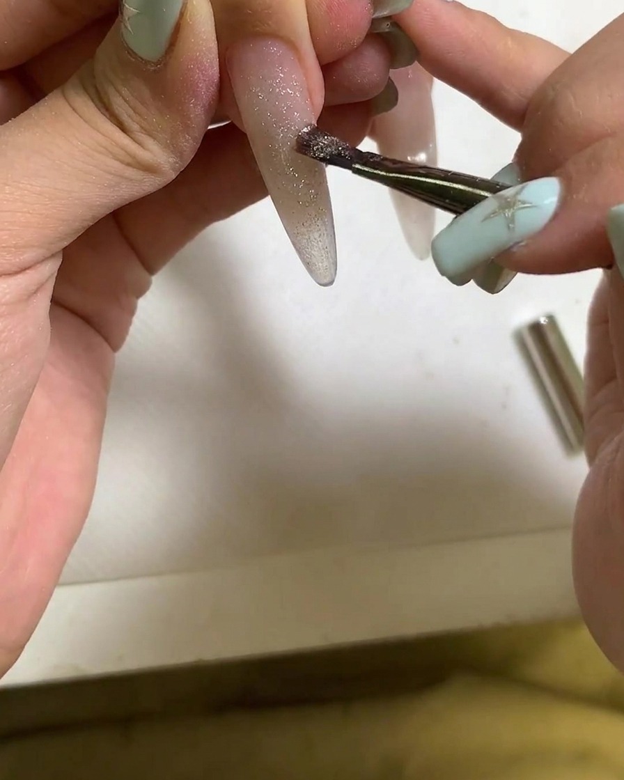 A brush applying cat eye gel polish onto an elongated fingernail.