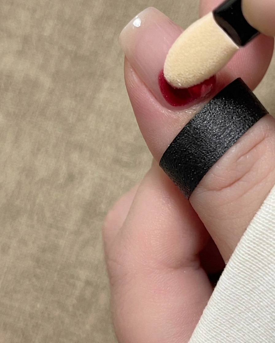 A sponge eyeshadow applicator dabbing red ink pigment at the base of a natural fingernail.